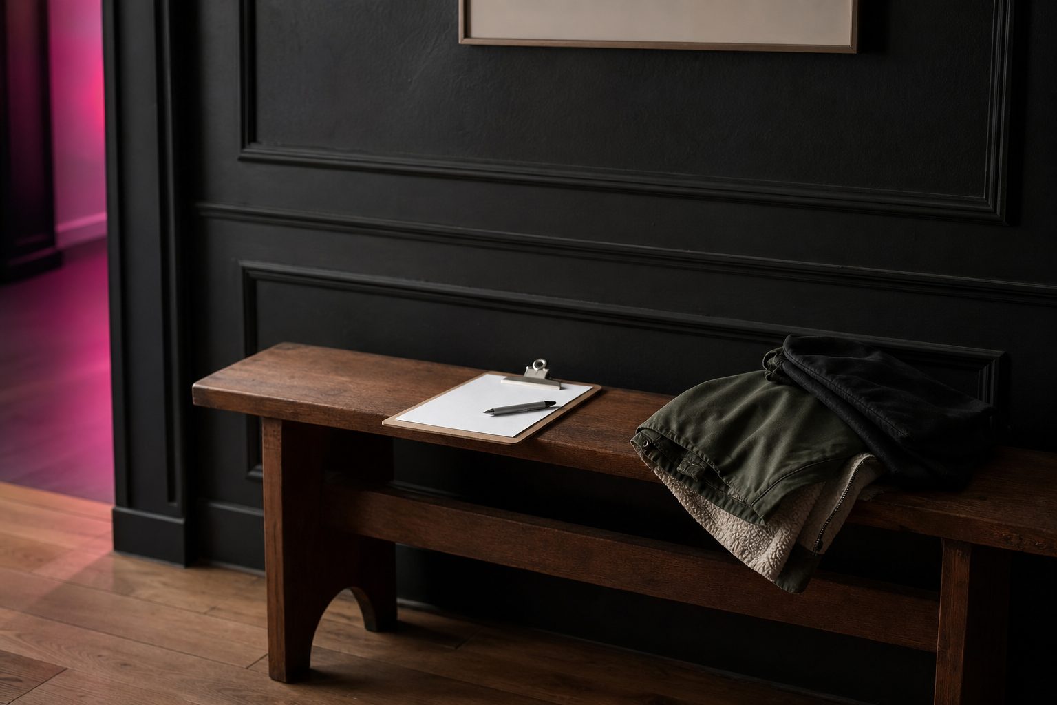 Studio bench with a clipboard, pen and a parent's coat against a dark paneled wall in soft light.