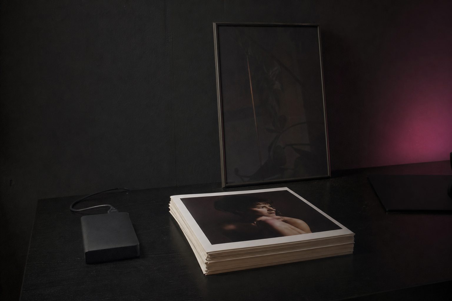 Stack of warm white portrait prints and an archive drive on a dark studio desk lit from the side.